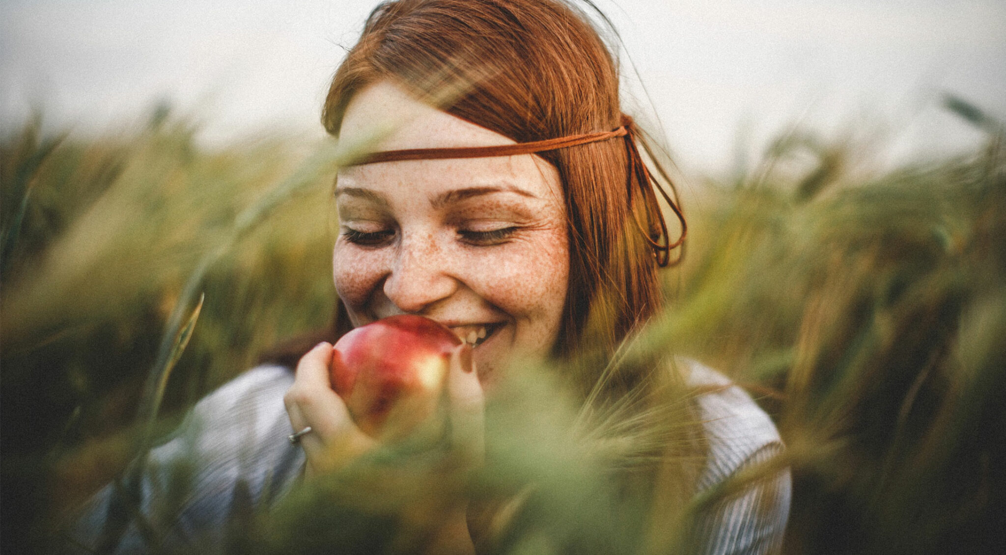 Frau mit roten Haaren steht in einem Feld und beißt in einen Apfel.