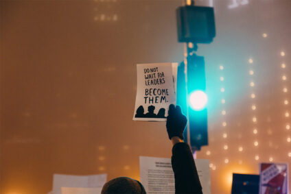 Jemand hält einen Zettel hoch mit der Aufschrift: "Don't wait for leaders. Become them."