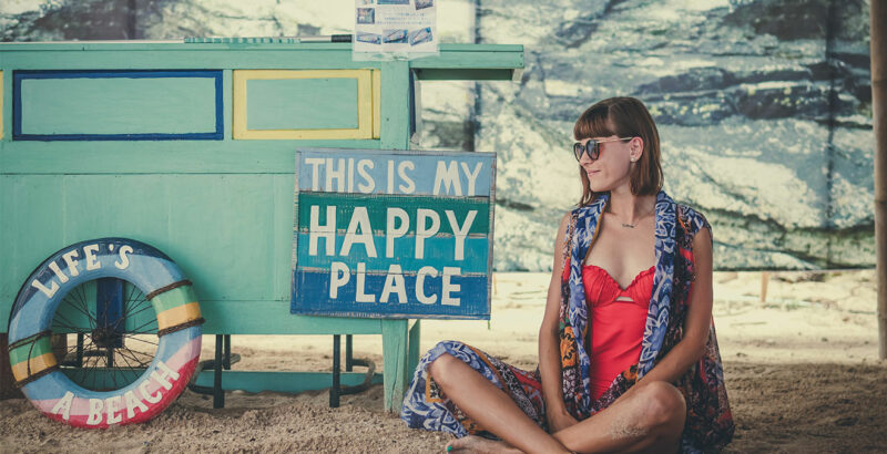 Eine Frau sitzt am Strand neben einem Holzschild mit der Aufschrift "This is my happy place".