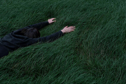Eine schwarz gekleidete Person liegt auf dem Bauch im tiefen Gras.