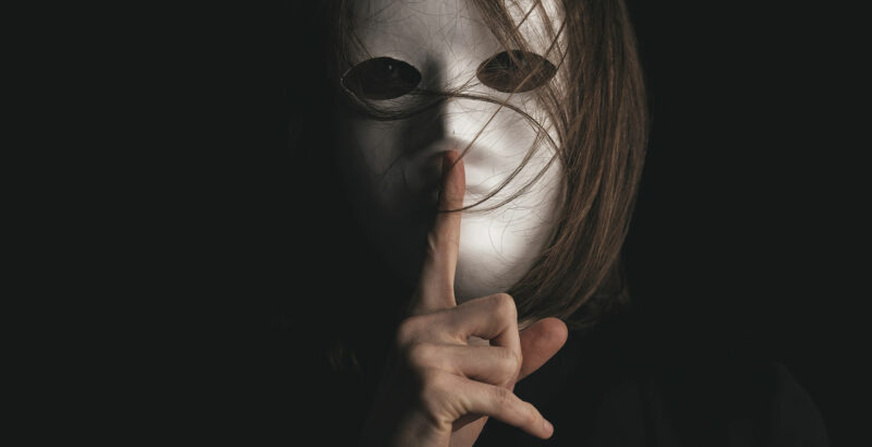 Mensch mit einer Maske hält den Zeigefinger vor den Mund - Schweigen