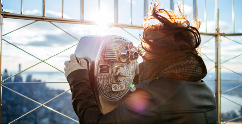 Eine winterlich angezogene Frau blickt mit wehenden Haaren durch ein Fernglas