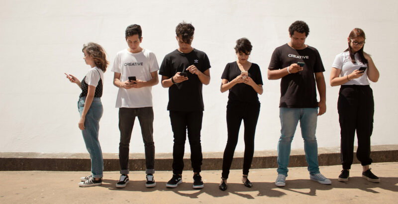 Eine Gruppe von sechs jungen Mensch steht vor weißem Hintergrund nebeneinander, alle mit dem Blick auf ein Smartphone