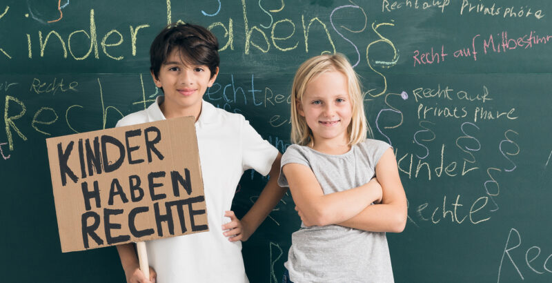 Ein Junge und ein Mädchen vor einer Schultafel mit einem Schild mit der Aufschrift "Kinder haben Rechte"