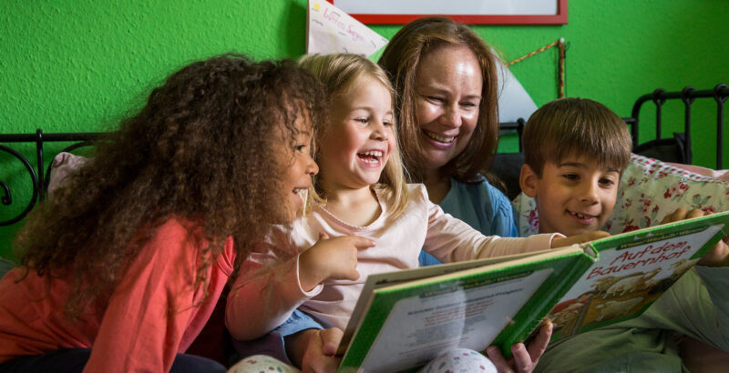 Eine Frau/Mutter liest drei Kindern ein Buch vor. Alle lachen