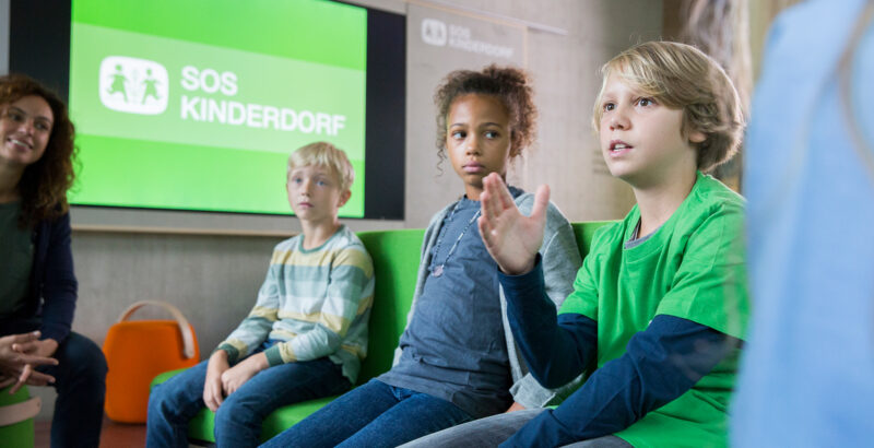 Drei Kinder sitzen und diskutieren vor einem Bildschirm mit dem Logo von SOS Kinderdorf
