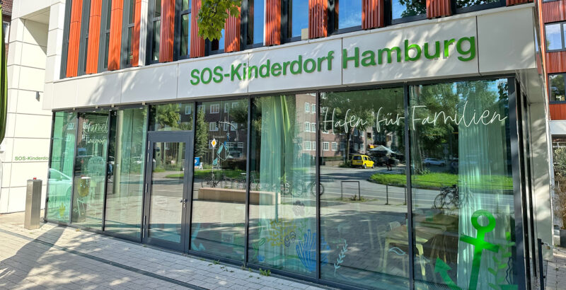 Glasfassade des SOS-Kinderdorfs Hamburg mit reflektierten Gebäuden und Schriftzug