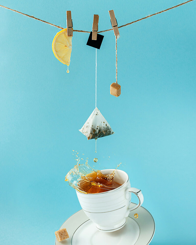 Eine weiße Teetasse mit Tee, über der mehrere Objekte hängen: eine Schaukel mit einem Teebeutel, eine Wippe und eine Lampe – surreal in hellblauer Umgebung.