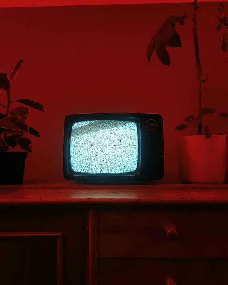 Ein alter Röhrenfernseher mit blauem Flimmern (Störsignal) steht in einem dunklen, rot beleuchteten Raum.