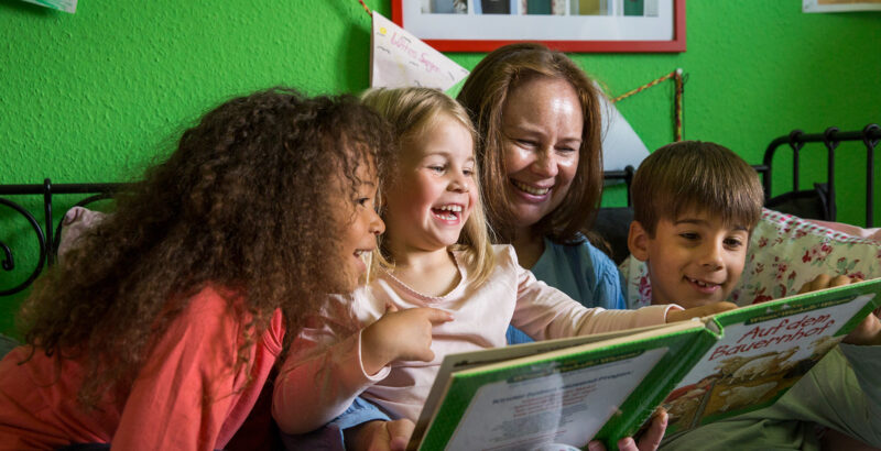 Eine Frau liest drei Kindern aus einem großen Buch vor.