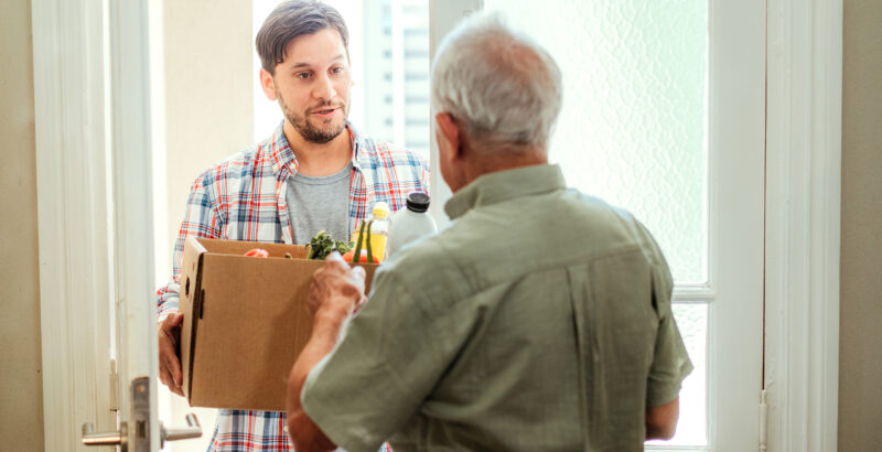 Junger Mann überreicht einem älteren Mann ein Paket mit Lebensmitteln und beide lächeln sich an