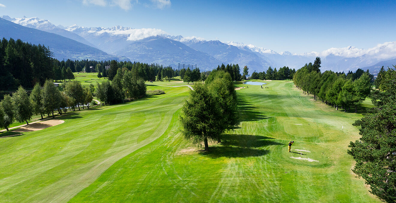 Golfplatz mit Bäumen vor Bergkulisse und Sonnenschein.