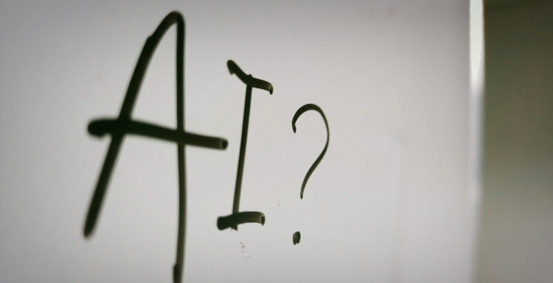 Schriftzug ‚AI?‘ auf weißem Hintergrund – Fragezeichen als Symbol für ethische und gesellschaftliche Fragen zur künstlichen Intelligenz.