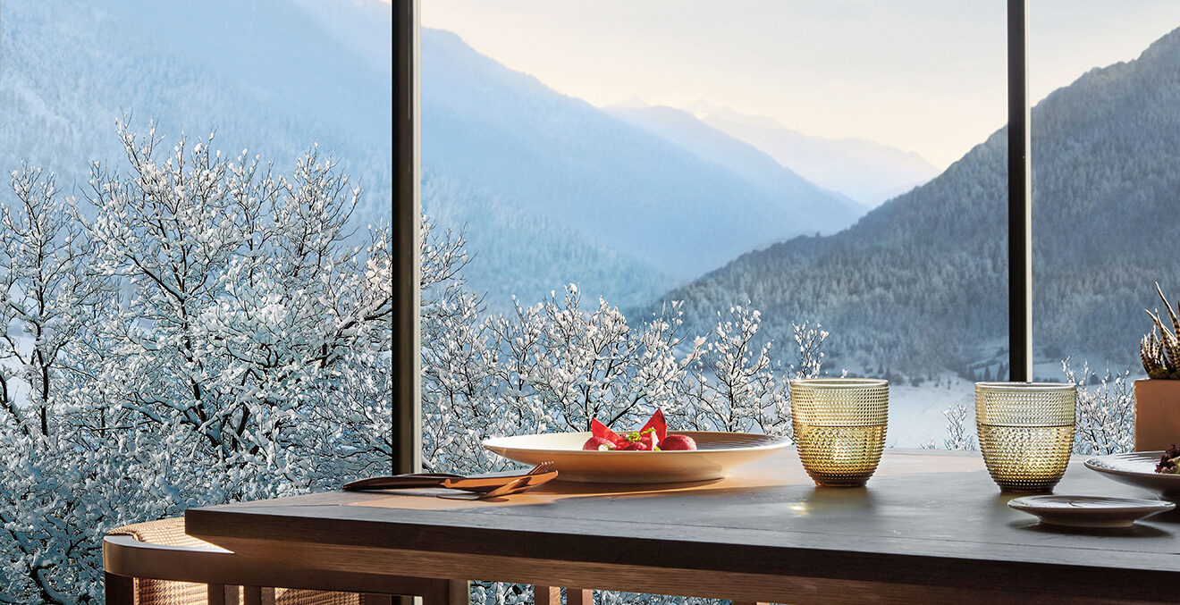 Tisch und Bank vor Glasfront mit Blick in die Berge.