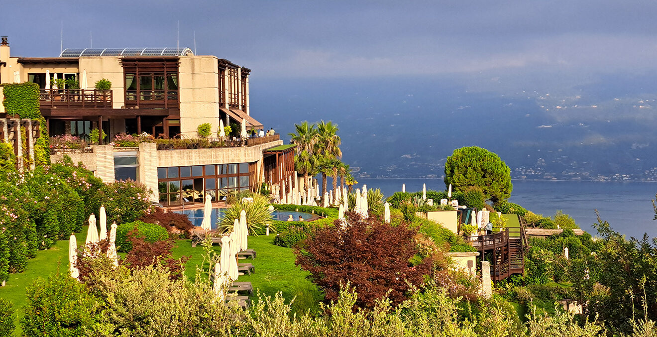 Panoramablick auf das Lefay Resort direkt am Ufer des Gardasees mit Palmen, Sonnenterrasse und Gebäuden in warmen Farben.