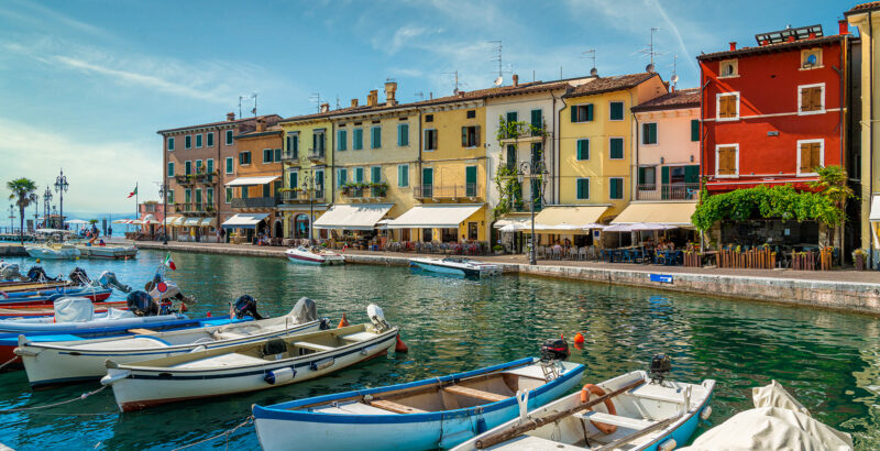 Gargnano - Steg mit Booten am Gardasee bei klarem Himmel, im Hintergrund sanfte Hügel und mediterrane Vegetation.“