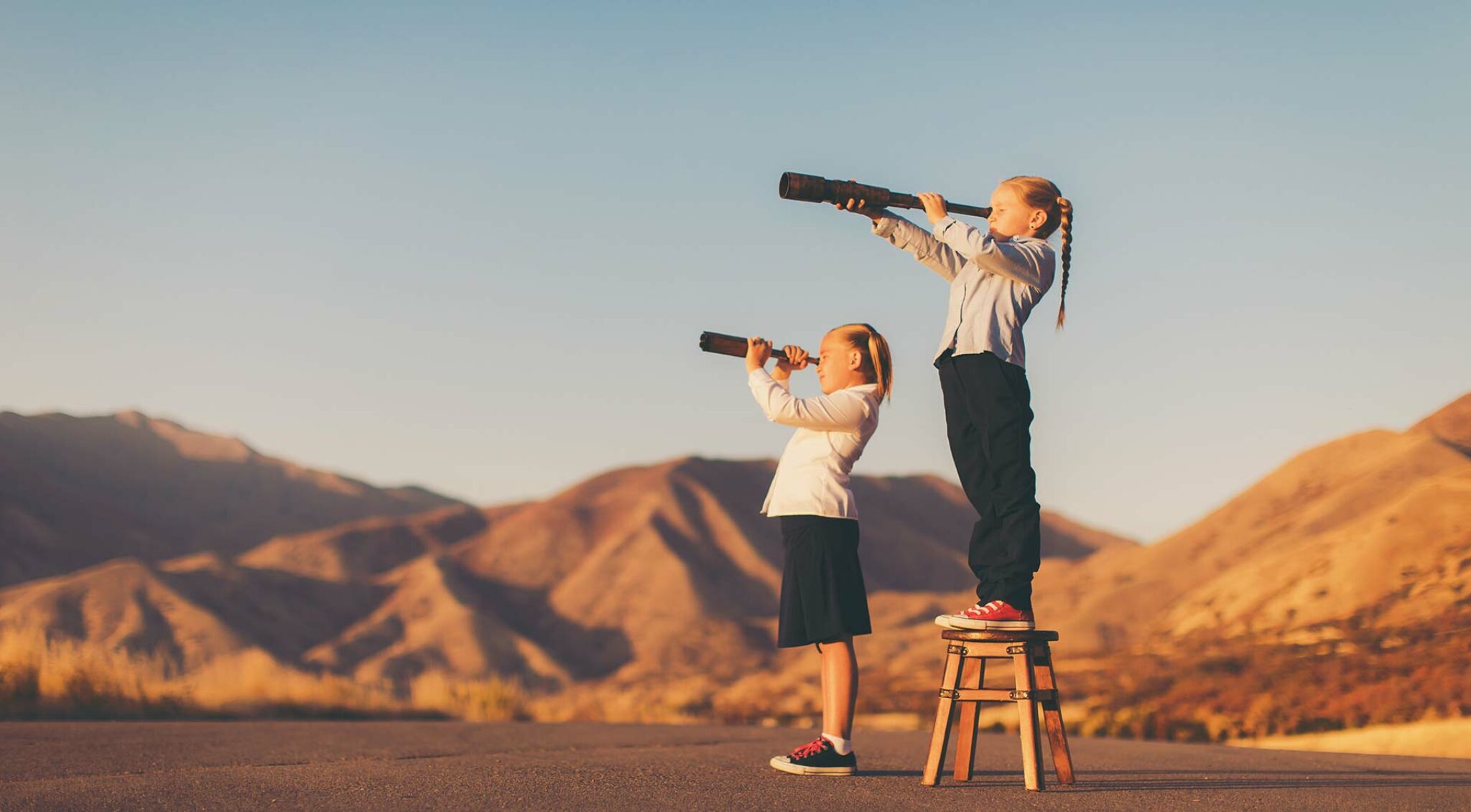 Zwei Mädchen stehen auf einem Hügel in einer hellen, weiten Berglandschaft und blicken jede durch ein Fernglas in die Ferne – Symbol für Orientierung, Zukunftssuche und Weitblick.