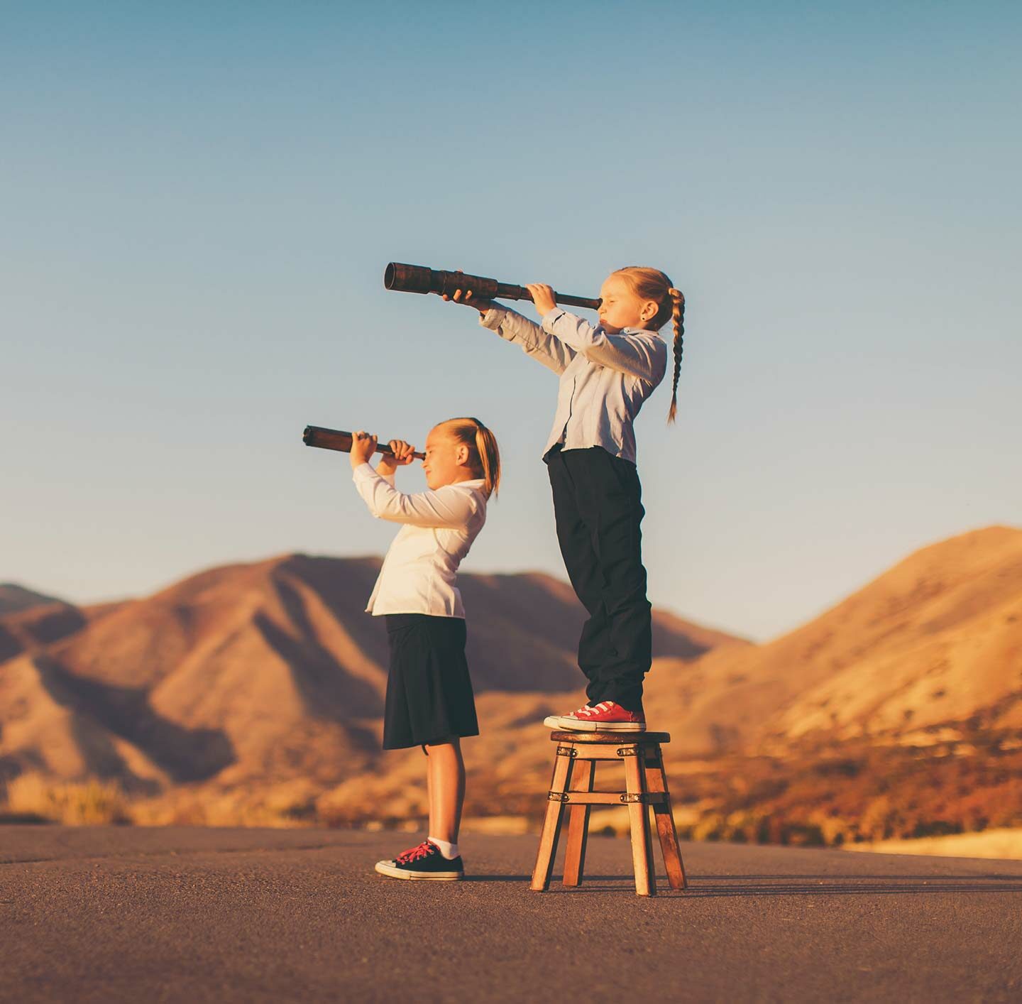 Zwei Mädchen stehen auf einem Hügel in einer hellen, weiten Berglandschaft und blicken jede durch ein Fernglas in die Ferne – Symbol für Orientierung, Zukunftssuche und Weitblick.