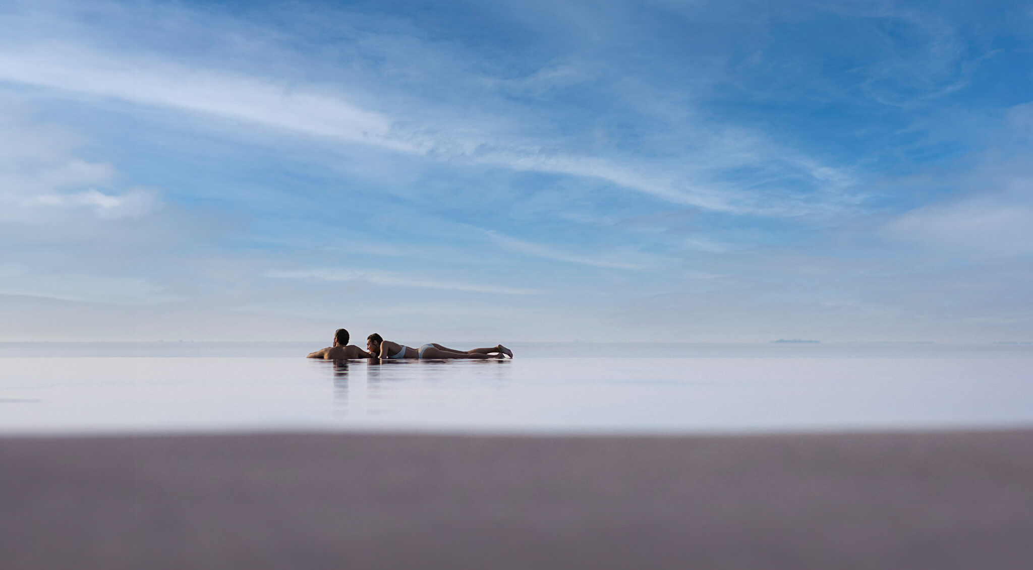 Weitläufige Luftaufnahme mehrerer kleiner Malediven-Inseln, umgeben von türkisem Wasser. Im Vordergrund zwei Personen im Infinitypool.