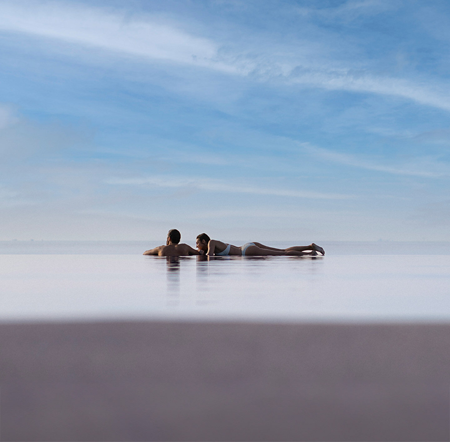 Weitläufige Luftaufnahme mehrerer kleiner Malediven-Inseln, umgeben von türkisem Wasser. Im Vordergrund zwei Personen im Infinitypool.