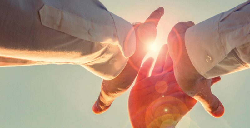 Drei Hände zeigen im warmen Gegenlicht zueinander. Die Szene vermittelt Vertrauen, Partnerschaft und Unterstützung in herausfordernden Zeiten.“