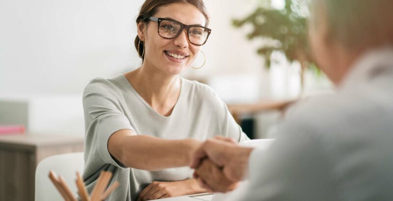 Eine Frau mit Brille lächelt und gibt einer anderen Person im Vordergrund die Hand; beide sitzen an einem Tisch in einer hellen, freundlichen Umgebung.