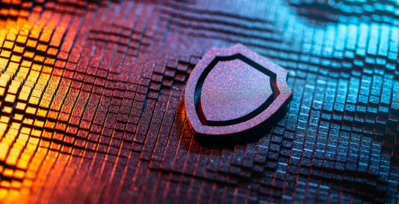 Nahaufnahme eines digitalen Schutzschild-Symbols, das auf einer blau-orange beleuchteten IT-Oberfläche liegt und Cybersicherheit darstellt.