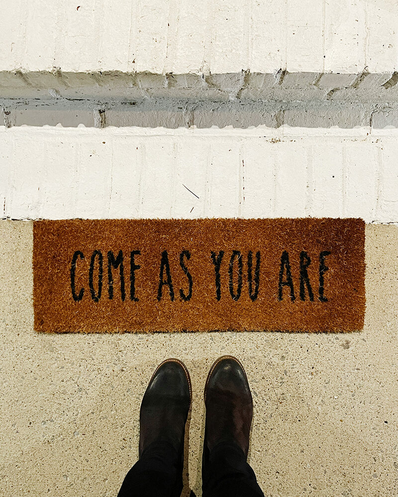 Fußmatte mit der Aufschrift „Come as you are“, davor zwei schwarze Schuhe.