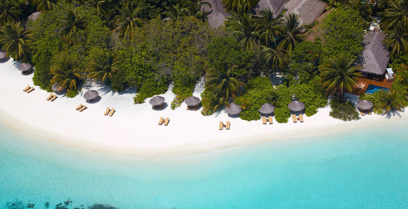 Blick von oben auf weißen Sandstrand mit Sonnenschirmen, Palmen und Holzhäuser.