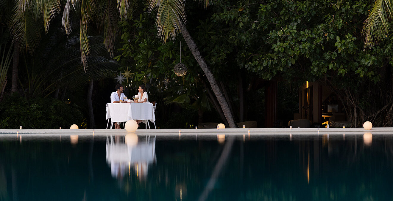 Weiss gedeckter Tisch mit zwei Personen unter Palmen am Wasser.