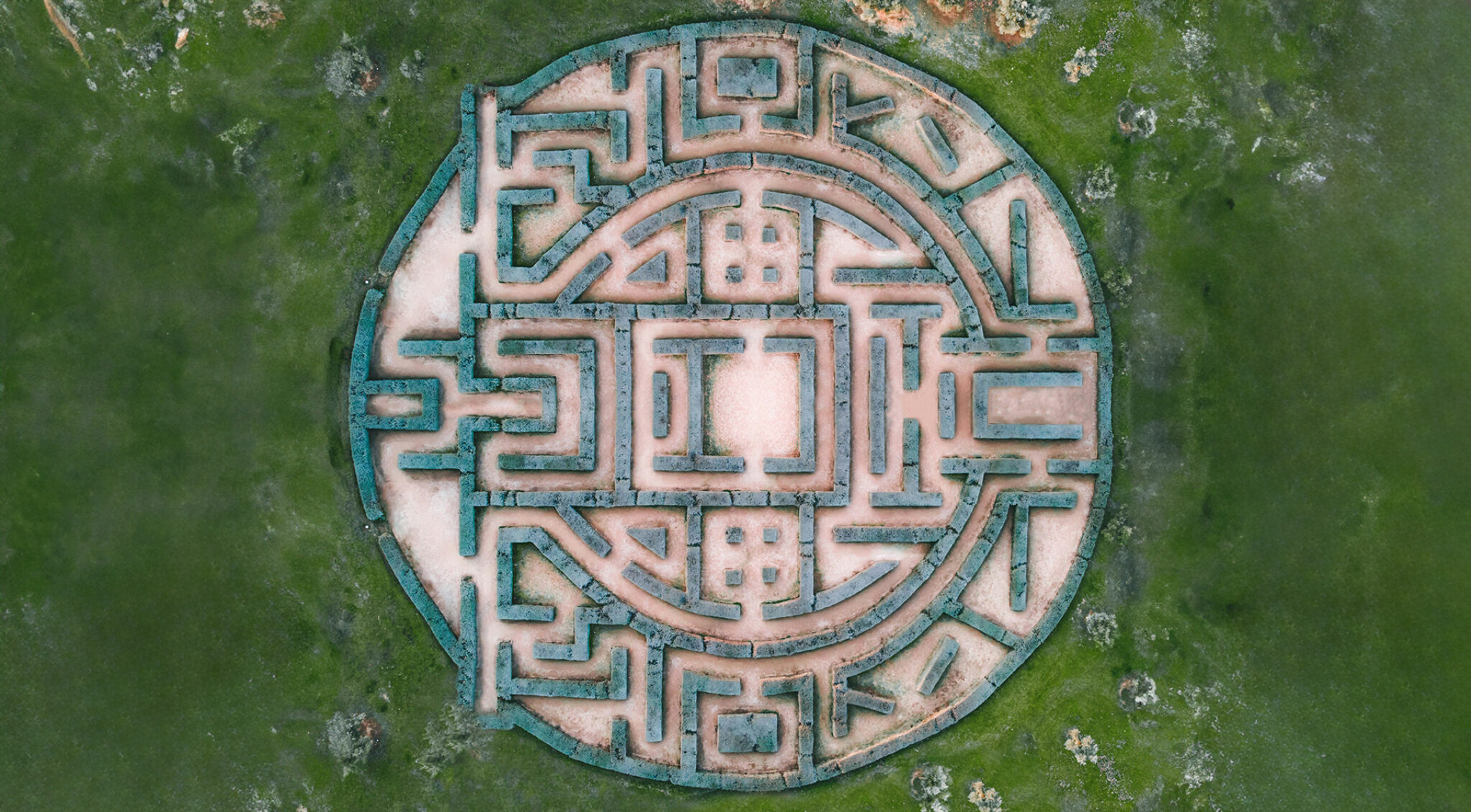 Die Aufnahme zeigt ein kreisförmiges Areal mit geometrischen Wegen und Strukturen, die an ein Mandala oder Labyrinth erinnern. Die umliegenden Bereiche sind grün bewachsen. Das Bild vermittelt eine Verbindung zwischen Struktur, Orientierung und Natur.