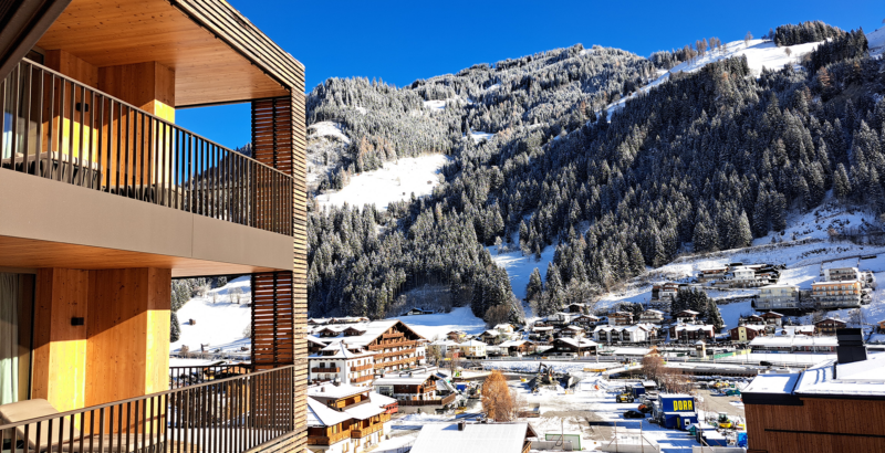 Modernes Hotelgebäude mit Holzfassade und Balkonen vor alpiner Bergkulisse.