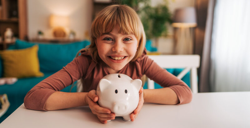 Ein Kind hält ein rosa Sparschwein auf einem Tisch und schaut lächelnd nach vorn.
