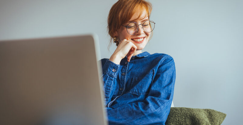 Junge Frau sitzt vor Laptop mit dem Blick nachdenklich zur Seite gerichtet.