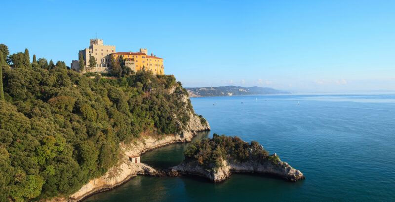 Küstenlandschaft mit einem monumentalen Bau auf einem bewaldeten Felsvorsprung und Blick auf das blaue Meer.