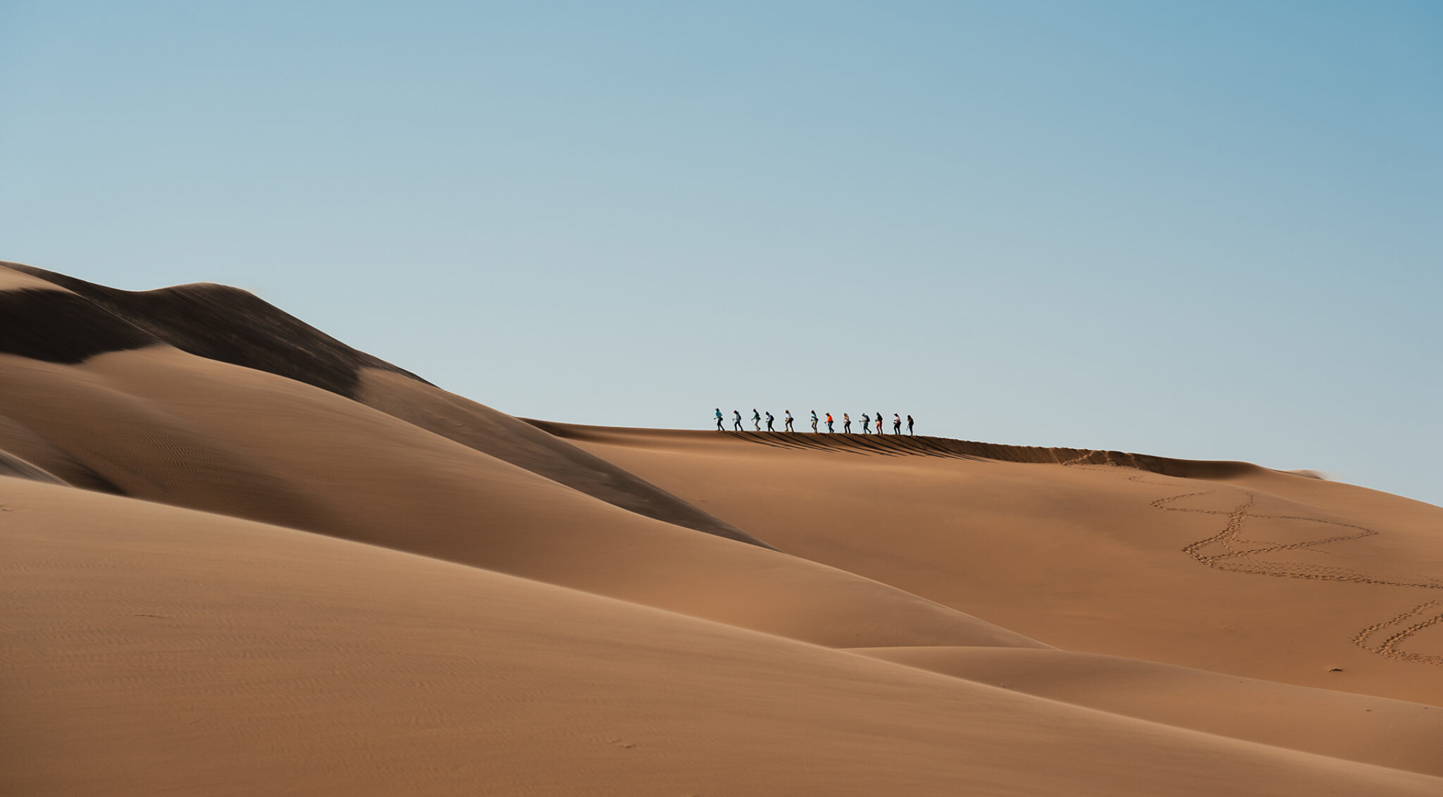 Weite Sanddünen der Namib-Wüste unter klarem Himmel, im Hintergrund eine kleine Karawane am Horizont.