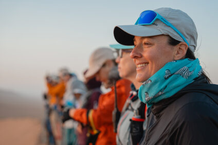 Mehrere Personen mit Kopfbedeckung und Ausrüstung bewegen sich durch die Wüste, teilweise unscharf dargestellt.