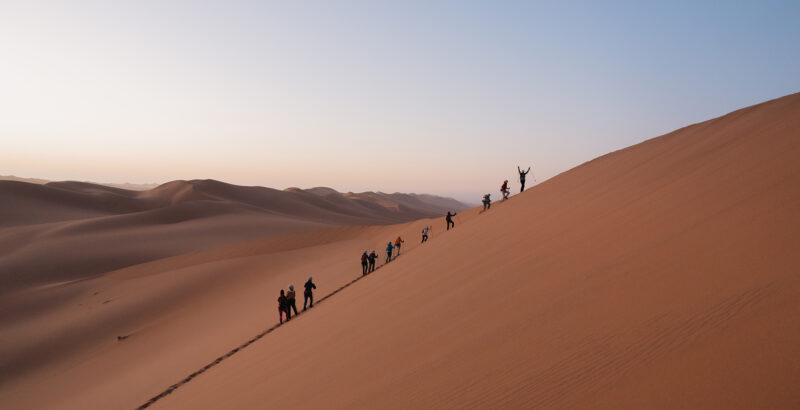 Gruppe von Menschen zieht in einer Linie über hohe Sanddünen, aufgenommen aus großer Distanz.