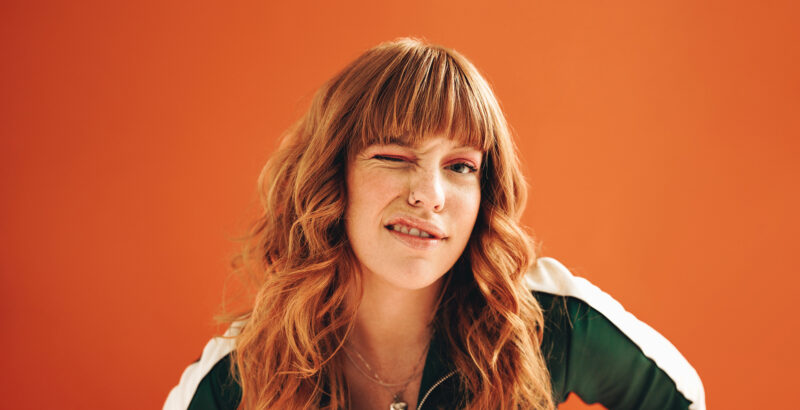 Porträt einer zwinkernden Frau mit lockigen Haaren vor orangefarbenem Hintergrund.