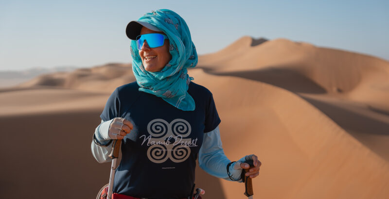 Frau mit Tuch über Kopf und Sonnenbrille steht in der Wüste und blickt über die Dünenlandschaft.