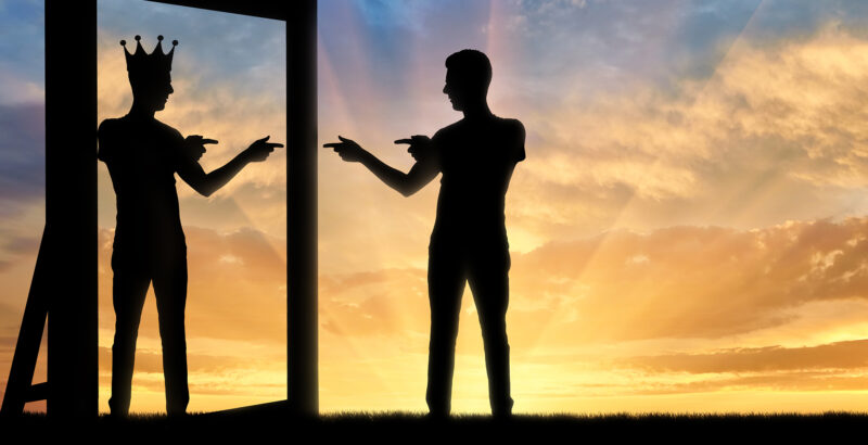 Zwei silhouettierte Personen stehen sich bei Sonnenuntergang gegenüber, getrennt durch einen Spiegel, wobei eine Silhouette eine Krone auf dem Kopf trägt.