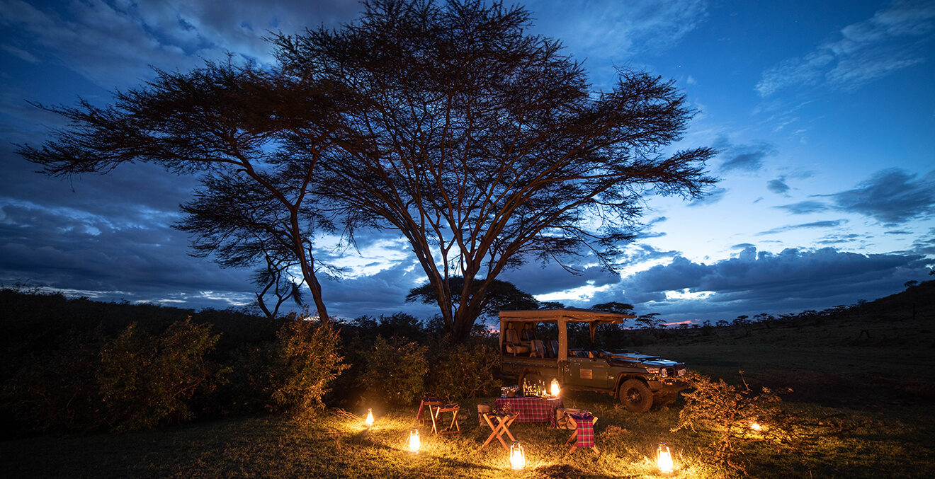 Panoramaaufnahme eines beleuchteten Camps in der Savanne unter blauem Abendhimmel.