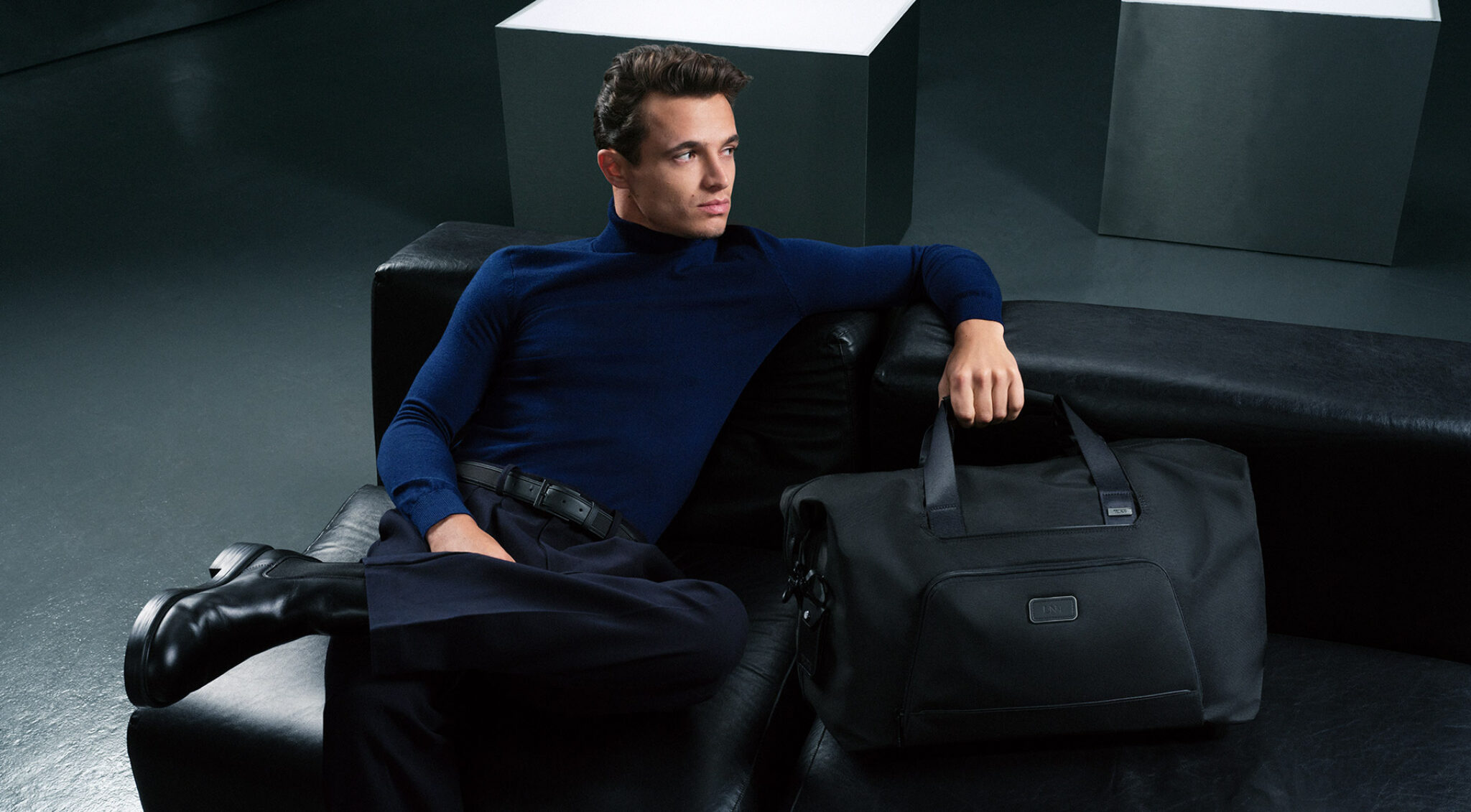 Mann in dunkelblauem Oberteil sitzt vor grauem Hintergrund neben einem großen schwarzen Rollkoffer; links im Vordergrund hält er eine kleine schwarze Tasche an einem Henkel.