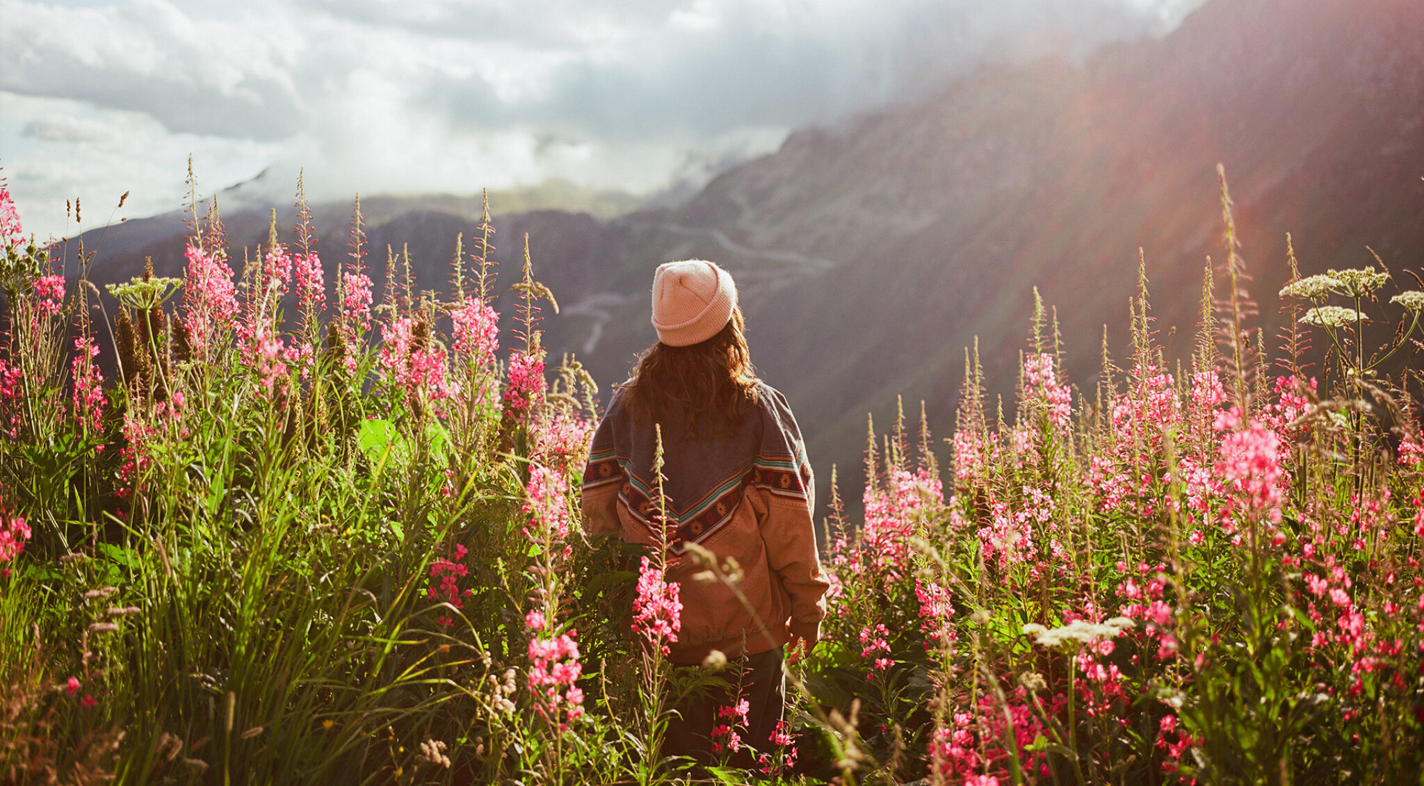 Person mit Mütze sitzt auf einer Wiese mit pinken Blumen und blickt über eine weite Berglandschaft.