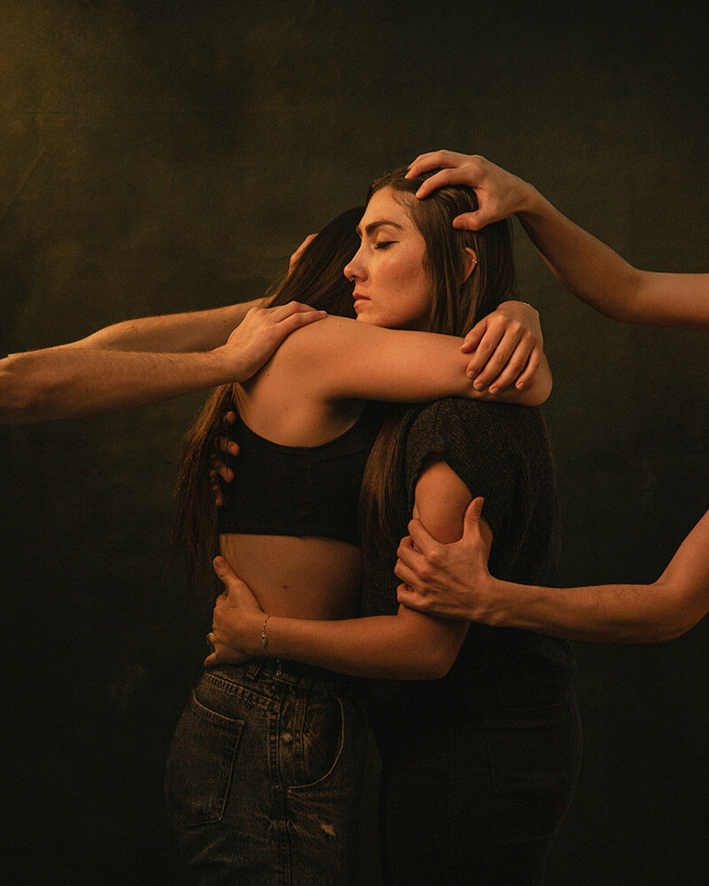 Zwei Frauen umarmen sich, während mehrere Hände ihre Schultern, Arme und den Kopf berühren.