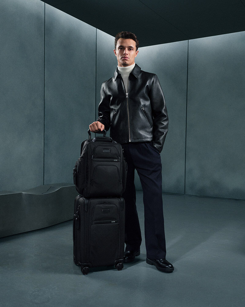 Person in dunkler Kleidung zieht einen großen schwarzen Rollkoffer, auf dem ein weiteres Gepäckstück steht, durch einen minimalistischen, grau beleuchteten Raum.