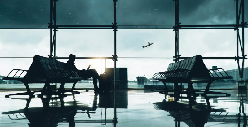 Silhouetten von Sitzreihen und Gepäckstücken vor großen Fenstern eines Flughafens; draußen ist ein Flugzeug zu sehen.