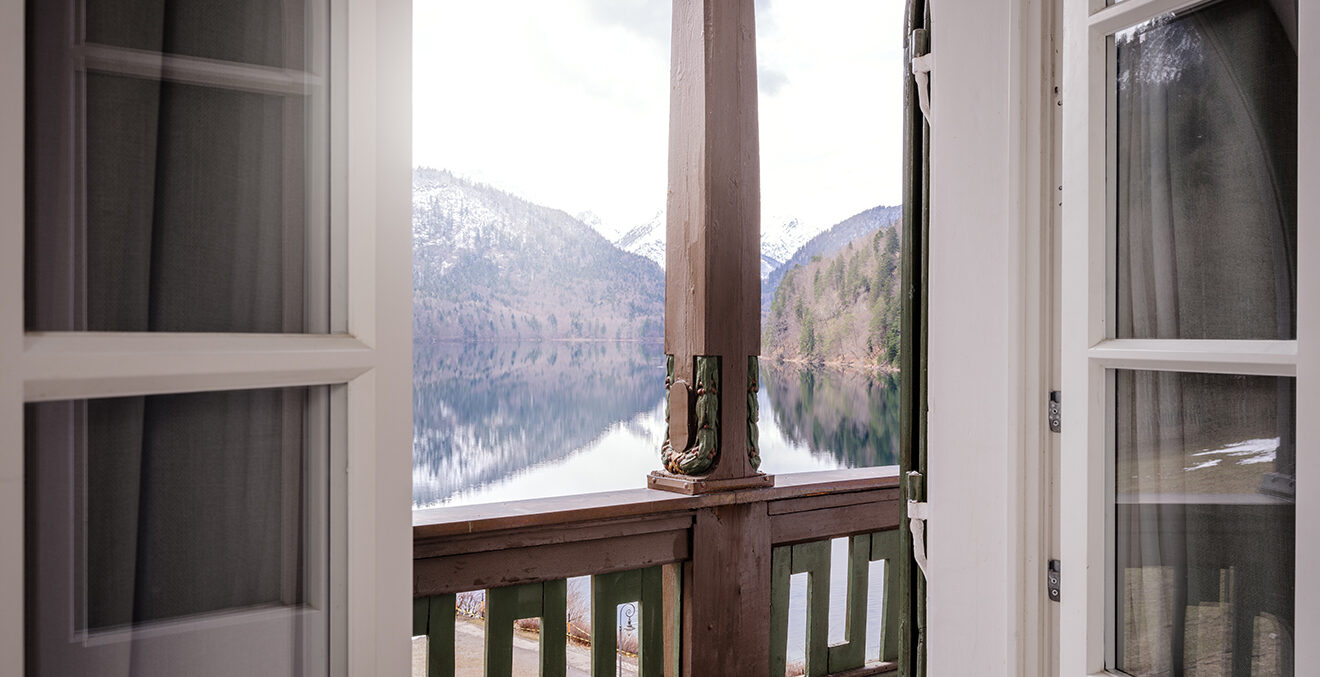 Blick durch eine offene Balkontür mit Geländer auf eine Landschaft mit See und Hügeln; Vorhänge rahmen die Öffnung.