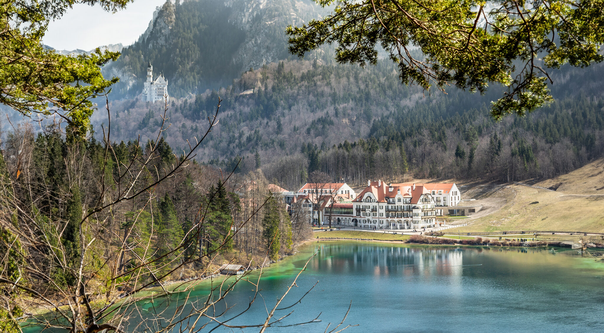 Blick über einen See auf eine weite Landschaft mit Wiesen, einzelnen Gebäuden und bewaldeten Hügeln im Hintergrund; im Vordergrund rahmen Äste mit grünen Blättern die Szene ein.