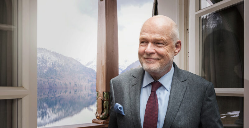 Hotelier Thomas Althoff sitzt vor einem Fenster; hinter ihm ist eine Landschaft mit See und Bergen sichtbar.